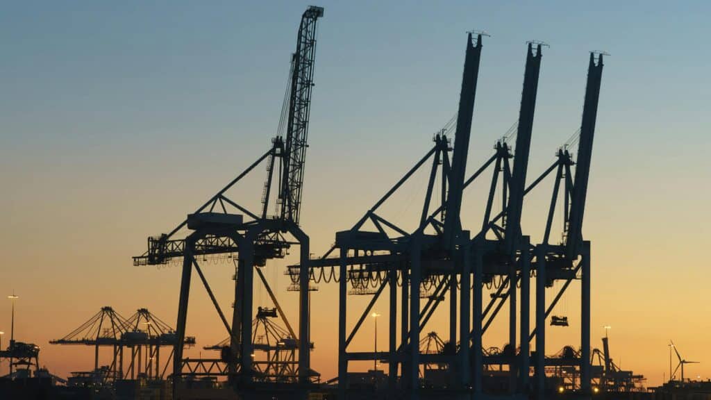 Silhouetted container cranes at an industrial shipping port during sunset, representing large-scale infrastructure and global logistics.