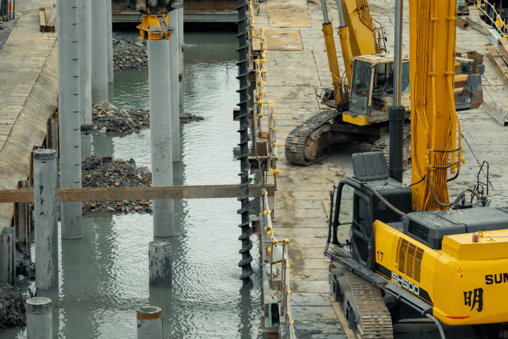 Excavators and large auger drill installing steel piles along a waterfront construction site for bridge foundation work.
