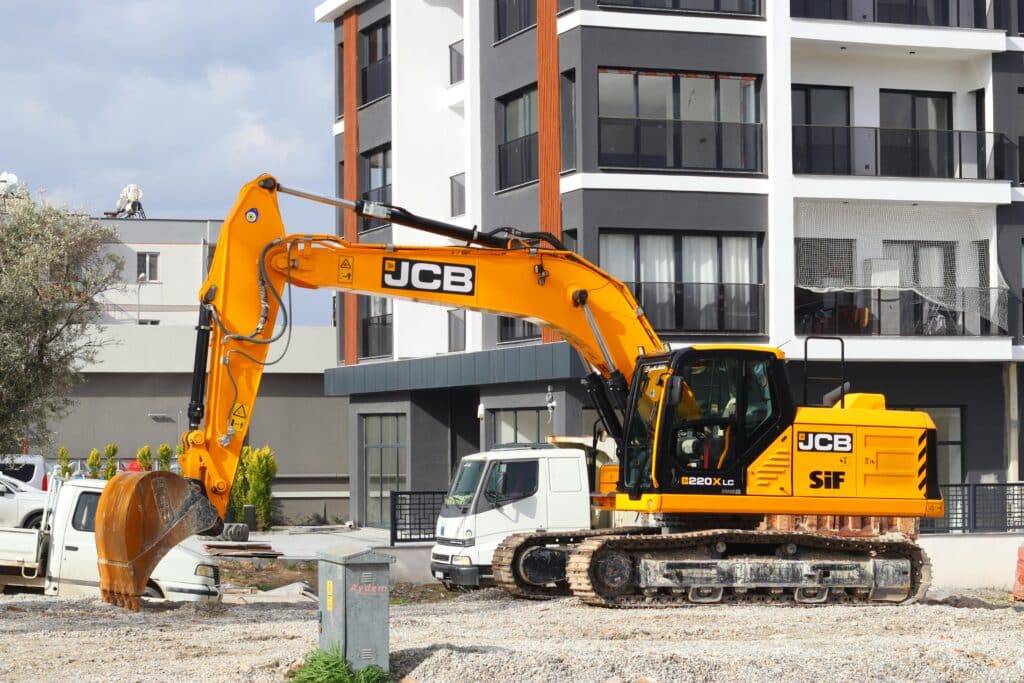 Yellow excavator operating at a modern apartment construction site with unfinished ground work and new residential buildings in the background