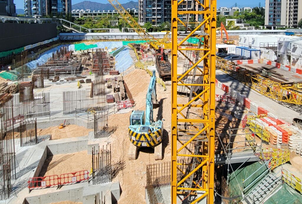 Commercial construction site with tower crane, excavator, and reinforced concrete foundations under active development.