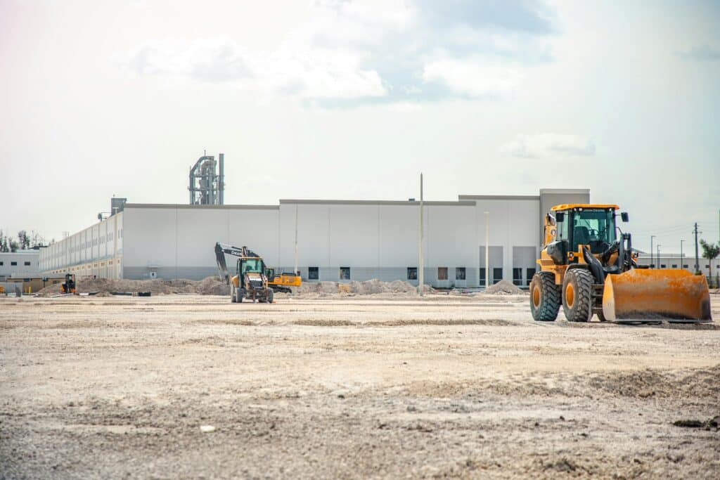 Front-end loader and excavator operating on a large dirt construction site in front of an industrial building.