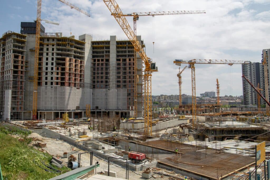 Urban high-rise buildings under construction with multiple tower cranes, concrete foundations, scaffolding, and workers on site