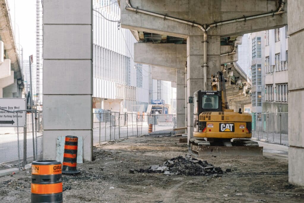Excavator grading dirt beneath elevated concrete transit structure at city construction site with traffic barrels and fencing