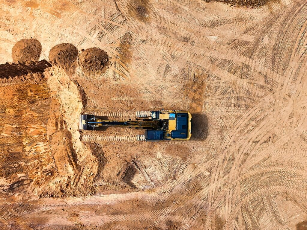 Aerial view of excavator digging trench in sandy soil with visible tire tracks at construction site