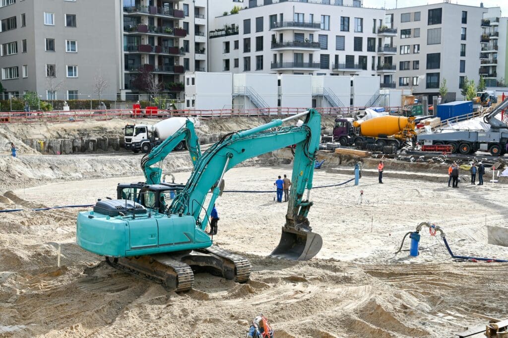 Hydraulic excavator operating at a large residential construction site with workers and concrete trucks in the background.