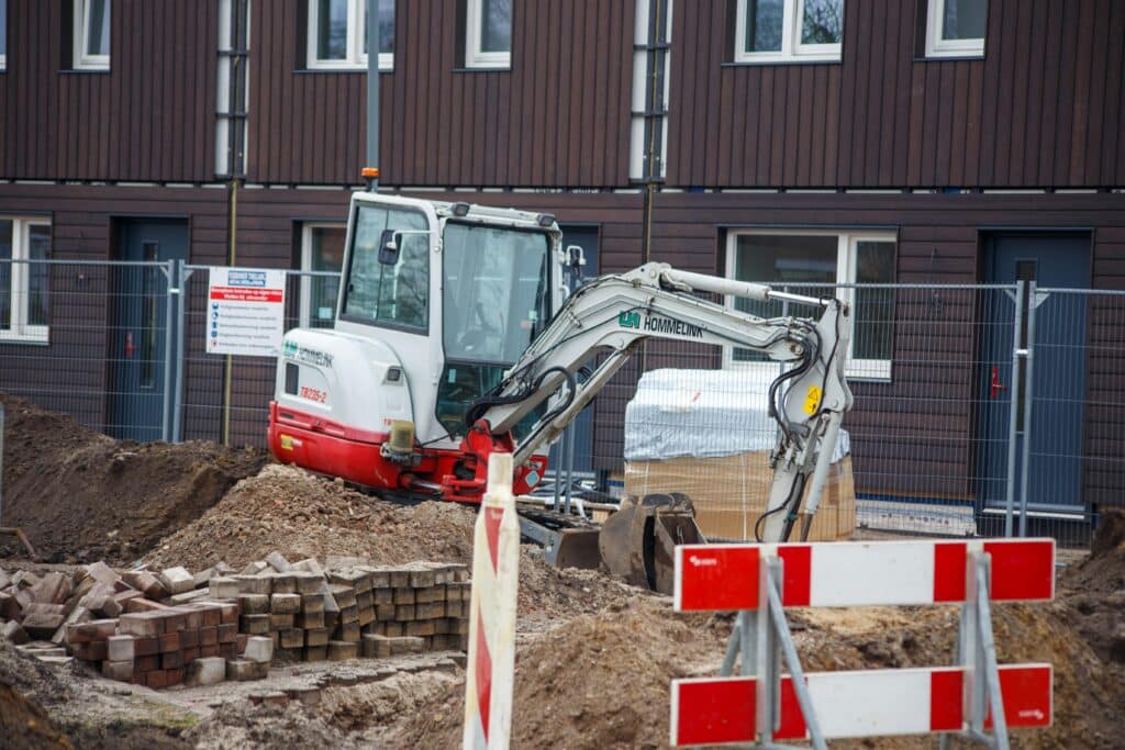 Excavator digging at a residential construction site with safety barriers and new housing in the background