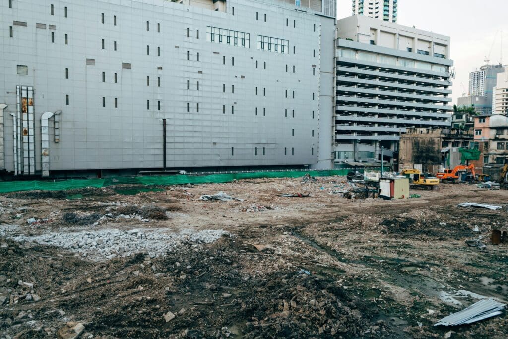 Large commercial construction site with heavy equipment beside a multi-story building and parking garage in an urban area